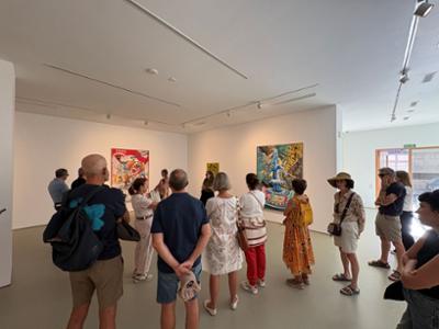 Visitantes recorren una exposición de arte contemporáneo en una galería, observando varias obras coloridas colgadas en paredes blancas durante una visita guiada en un espacio cultural moderno.