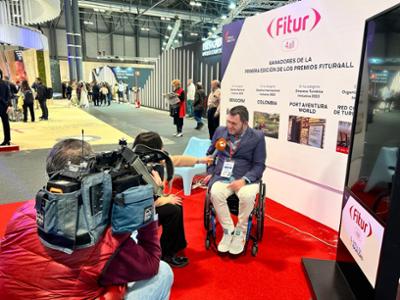 Unos periodistas entrevistan a una persona sentada en una silla de ruedas delante de un cartel de FITUR 4all