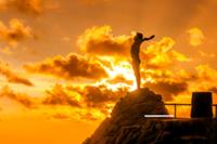 Persona encima de una montaña con los brazos en cruz en un amanecer con el sol detrás Fitur 2026