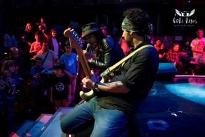 Guitarrista de rodillas tocando y detrás el cantante con un monton de personas y niños en el público en Rock en Familia