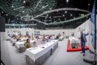 Conferencia Salón Peñín en un auditorio amplio, con mesas dispuestas en filas donde varias personas están sentadas escuchando a un presentador en el escenario. El techo tiene una estructura de entramado metálico y una iluminación tenue. El presentador está de pie junto a una pantalla grande, gesticulando mientras habla