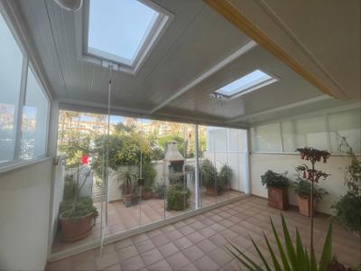 Terraza acristalada con dos claraboyas, suelo de baldosas, plantas ornamentales y una barbacoa de obra en un patio interior iluminado por luz natural.
