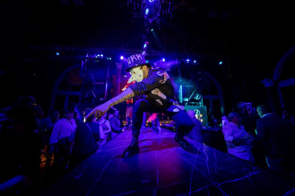 personaje de WAH con máscara blanca arrodillado señalando al público